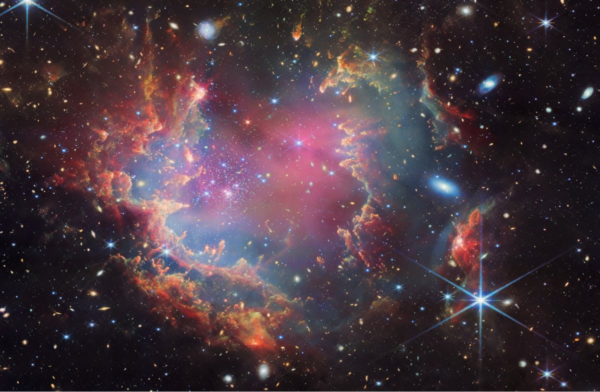 A star cluster is shown inside a large nebula of many-coloured gas and dust. The material forms dark ridges and peaks of gas and dust surrounding the cluster, lit on the inner side, while layers of diffuse, translucent clouds blanket over them. Around and within the gas, a huge number of distant galaxies can be seen, some quite large, as well as a few stars nearer to us which are very large and bright.