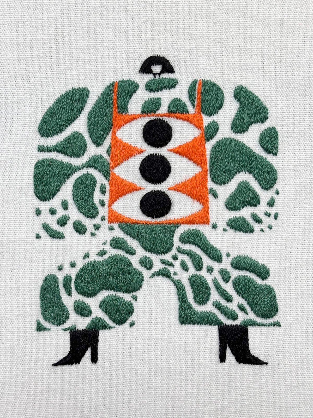 Embroidered figure in green leaf-patterned outfit with orange center panel containing three black circles