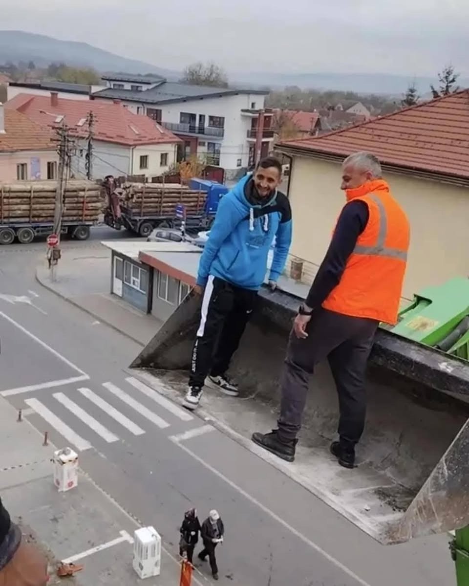 two guys standing on a front loader raised in the air; it looks like they are giants compared to all around them