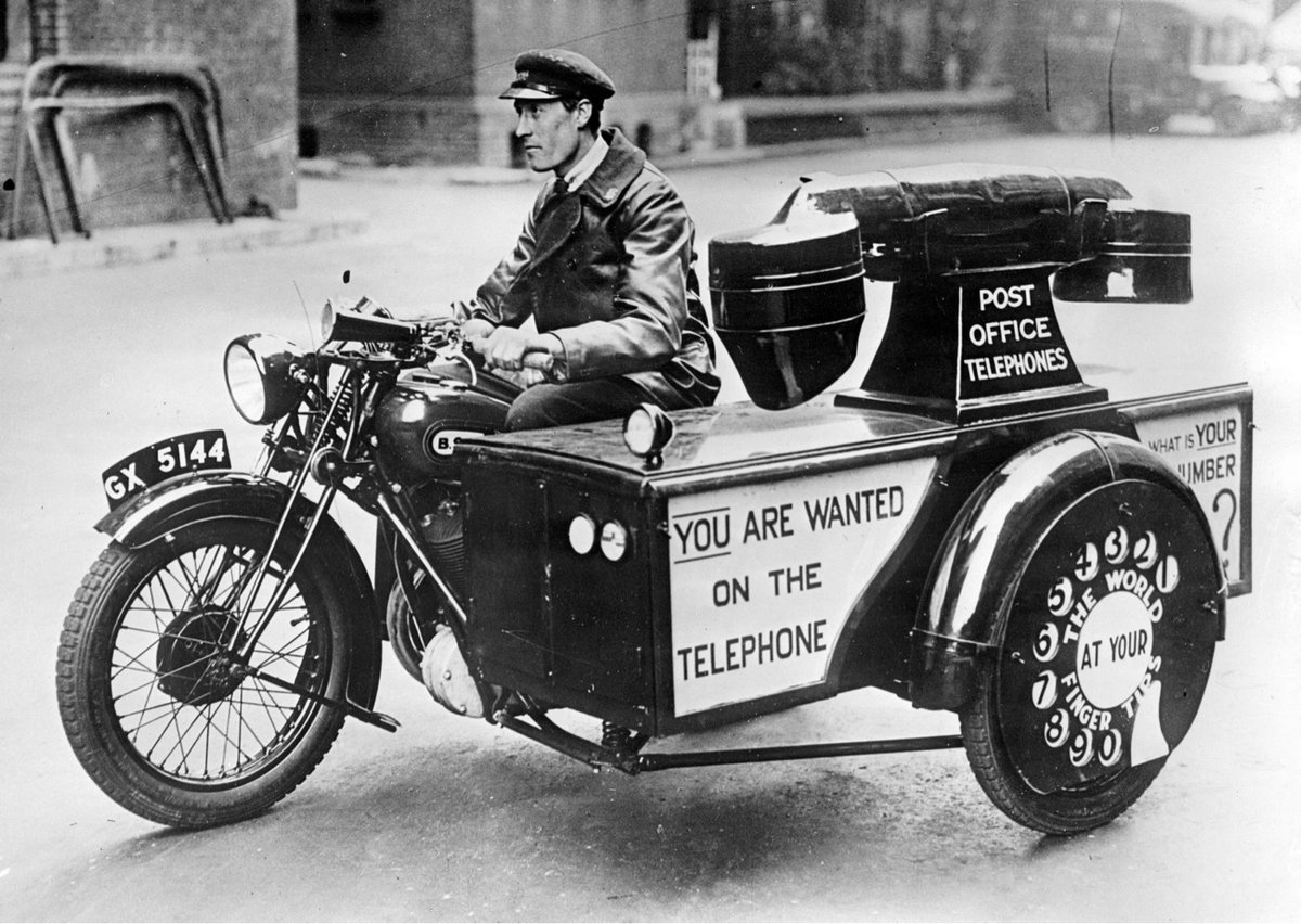 a black & white photo of a man driving a motorcycle with a sidecar shaped like a giant telephone