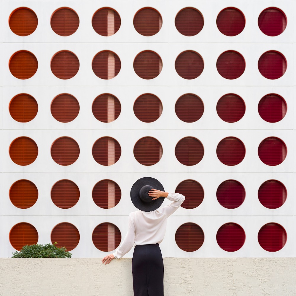 a woman stands in front of a wall covered in holes and her hat appears to be one of the holes